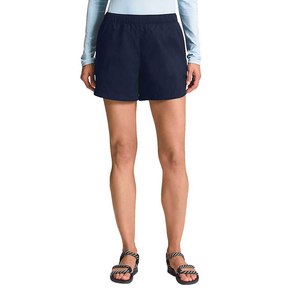 A person wearing a light blue long-sleeve shirt, The North Face Womens Class V Short in dark navy, and black sandals with white striped straps stands against a plain white background.