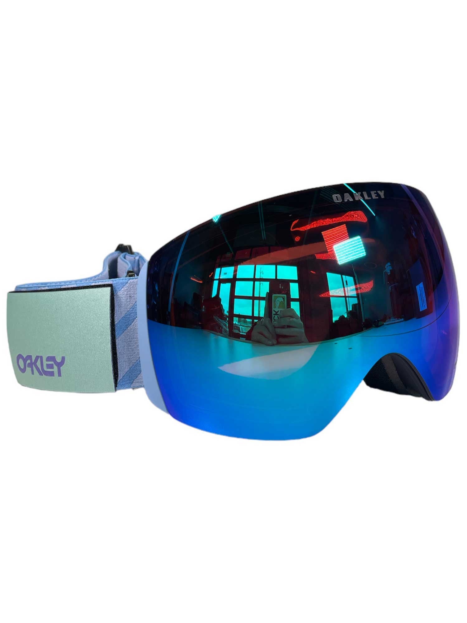Blue mirrored Oakley Flight Deck L Goggle featuring Prizm lens technology, a wide lens, and light blue strap with purple Oakley logo. The reflective lens displays a room lit by red neon and someone taking a photo.