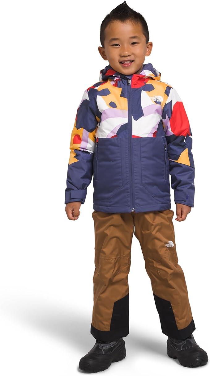 A smiling young child wears The North Face Toddlers Freedom Insulated Jacket with blue, red, yellow, and white shapes, brown pants with black knee patches, and black shoes, standing against a white background.