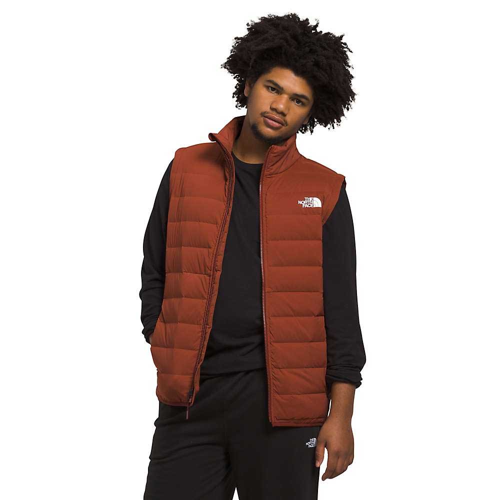 A man with curly hair wears The North Face Mens Belleview Stretch Down Vest in rust over a black long-sleeve shirt and black pants, standing against a white background with a relaxed expression.