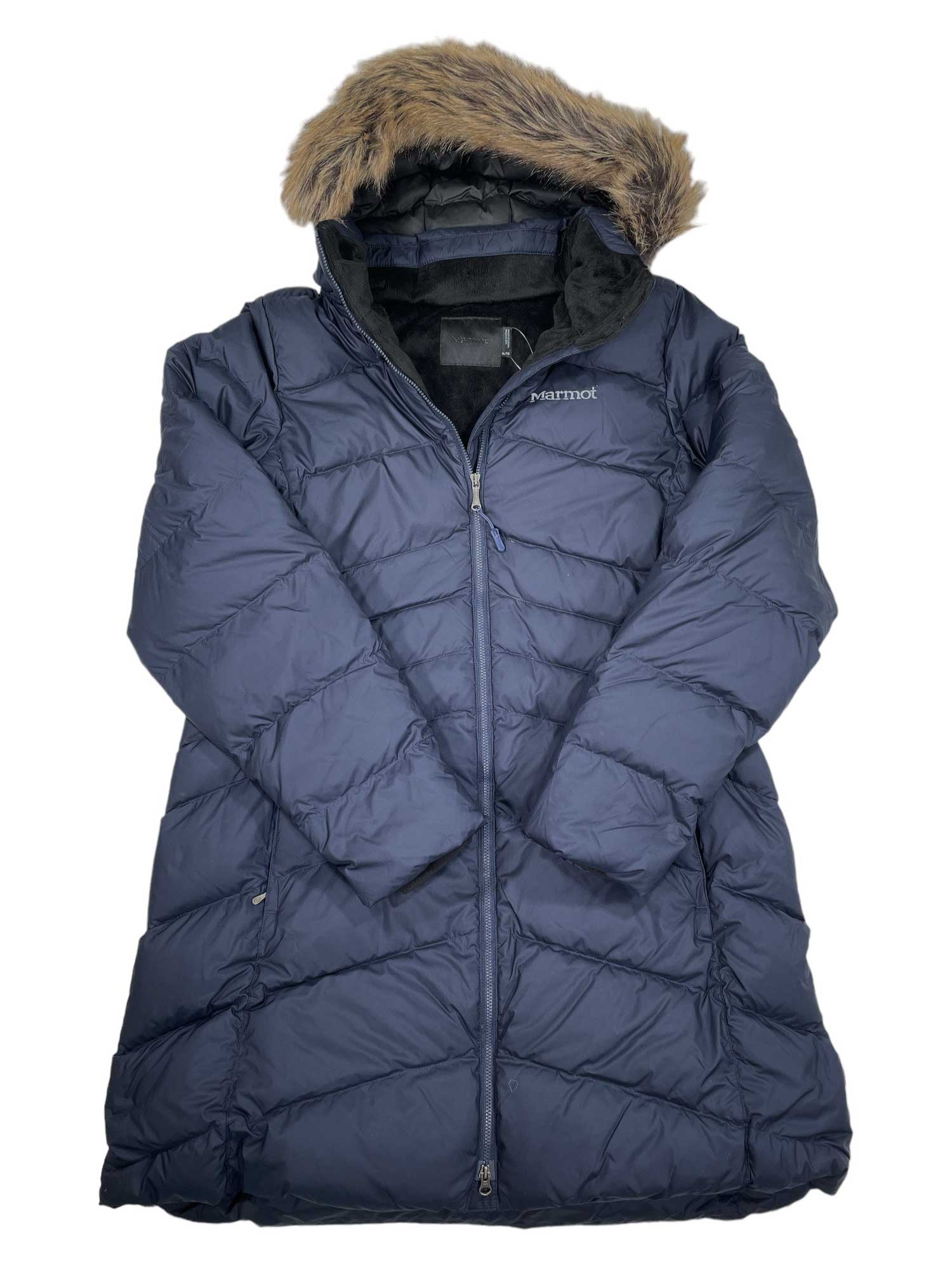 The Marmot Womens Montreal Down Coat is a navy quilted winter coat with a faux fur-lined hood, full zip, chest logo, and insulated water-resistant design. Laid flat, it highlights its long length and stylish warmth.