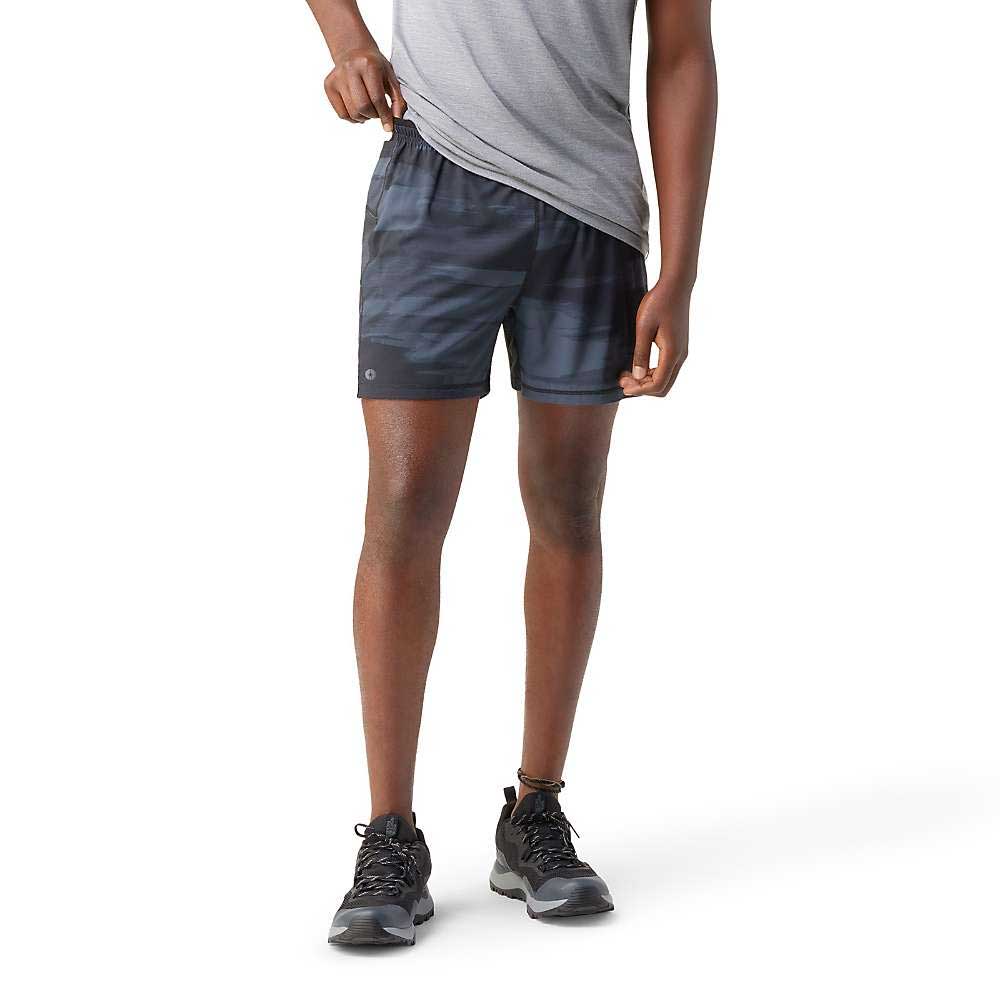 A person in a gray t-shirt and Smartwool Mens Active Lined 5 Inch Short stands against a white background, adjusting the shorts’ waistband with one hand. They wear black sneakers, and their face is not shown.