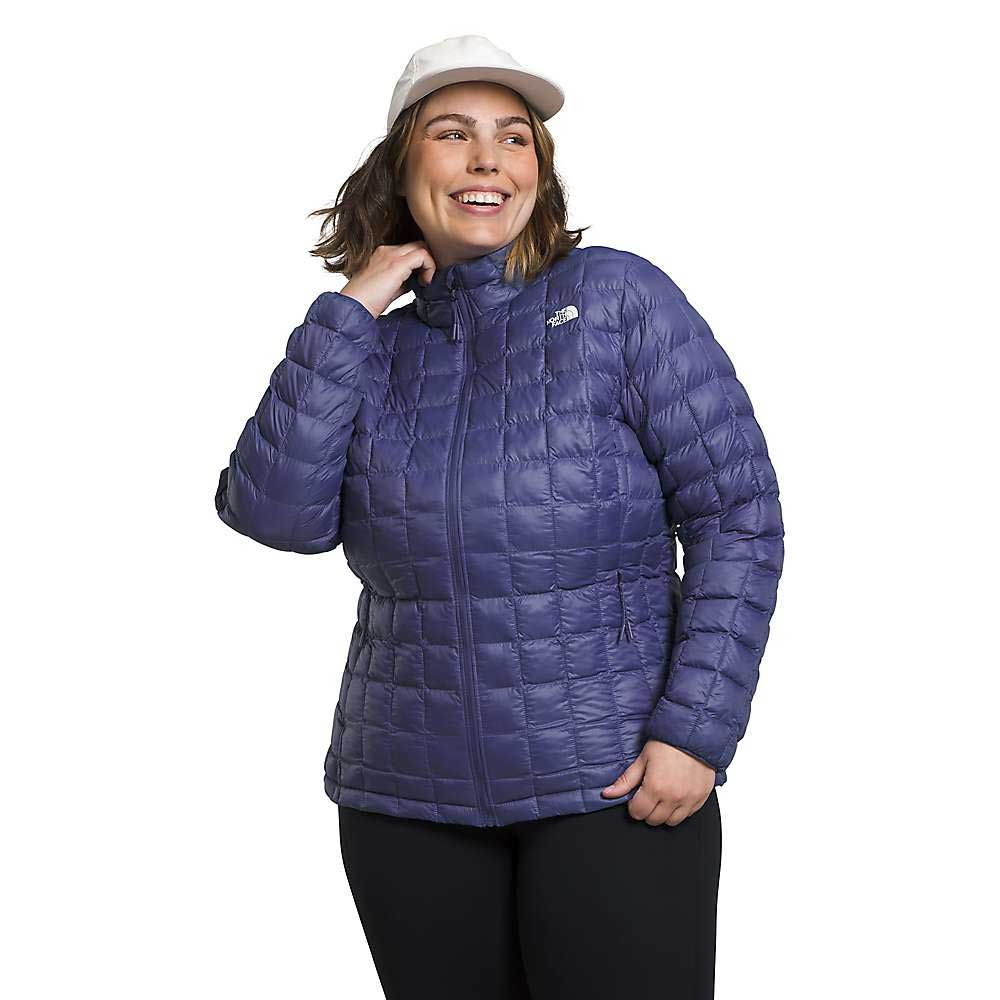 A smiling woman in The North Face Womens Plus ThermoBall Eco 2.0 Jacket, a white cap, and black pants stands with one hand on her collar against a plain white background.