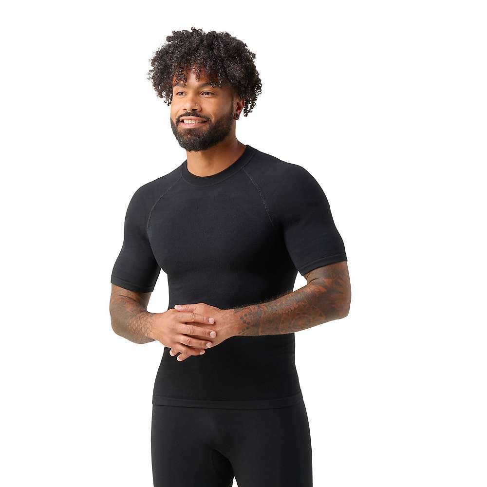 A man with curly hair and a beard smiles while wearing the Smartwool Men's Intraknit Active SS Top and matching pants, standing against a plain white background.