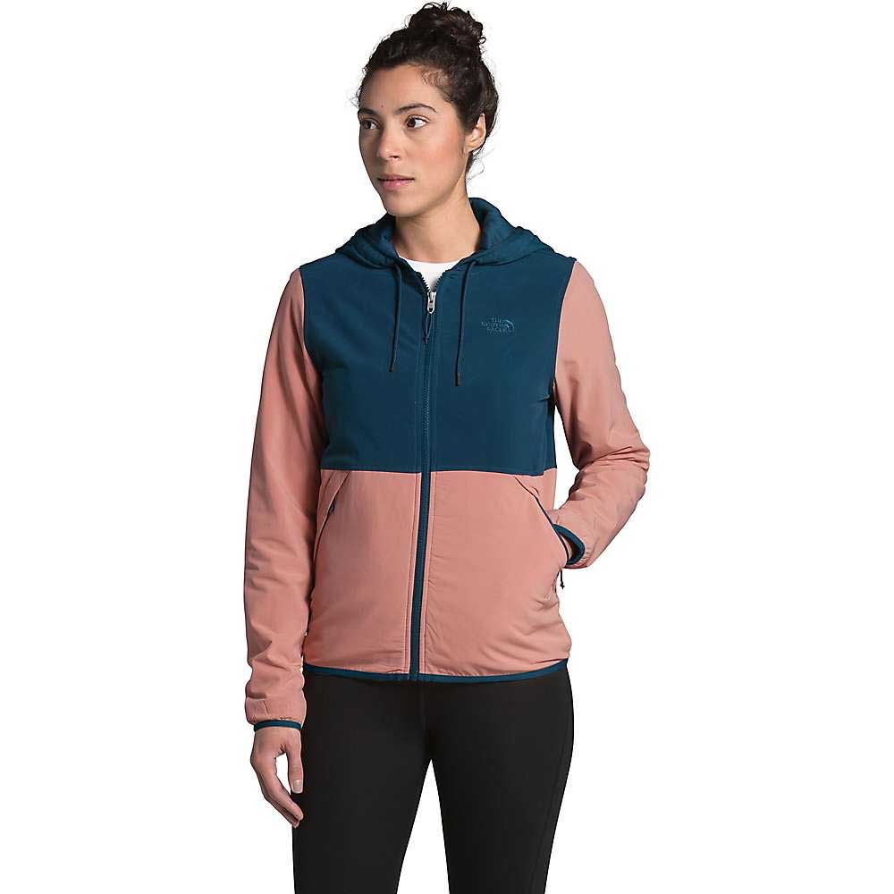 A woman with dark hair pulled back wears the Women's Mountain Sweatshirt Hoodie 3.0 by The North Face in dark teal and dusty pink color blocks, paired with black leggings. She stands looking to the side with one hand in her pocket.