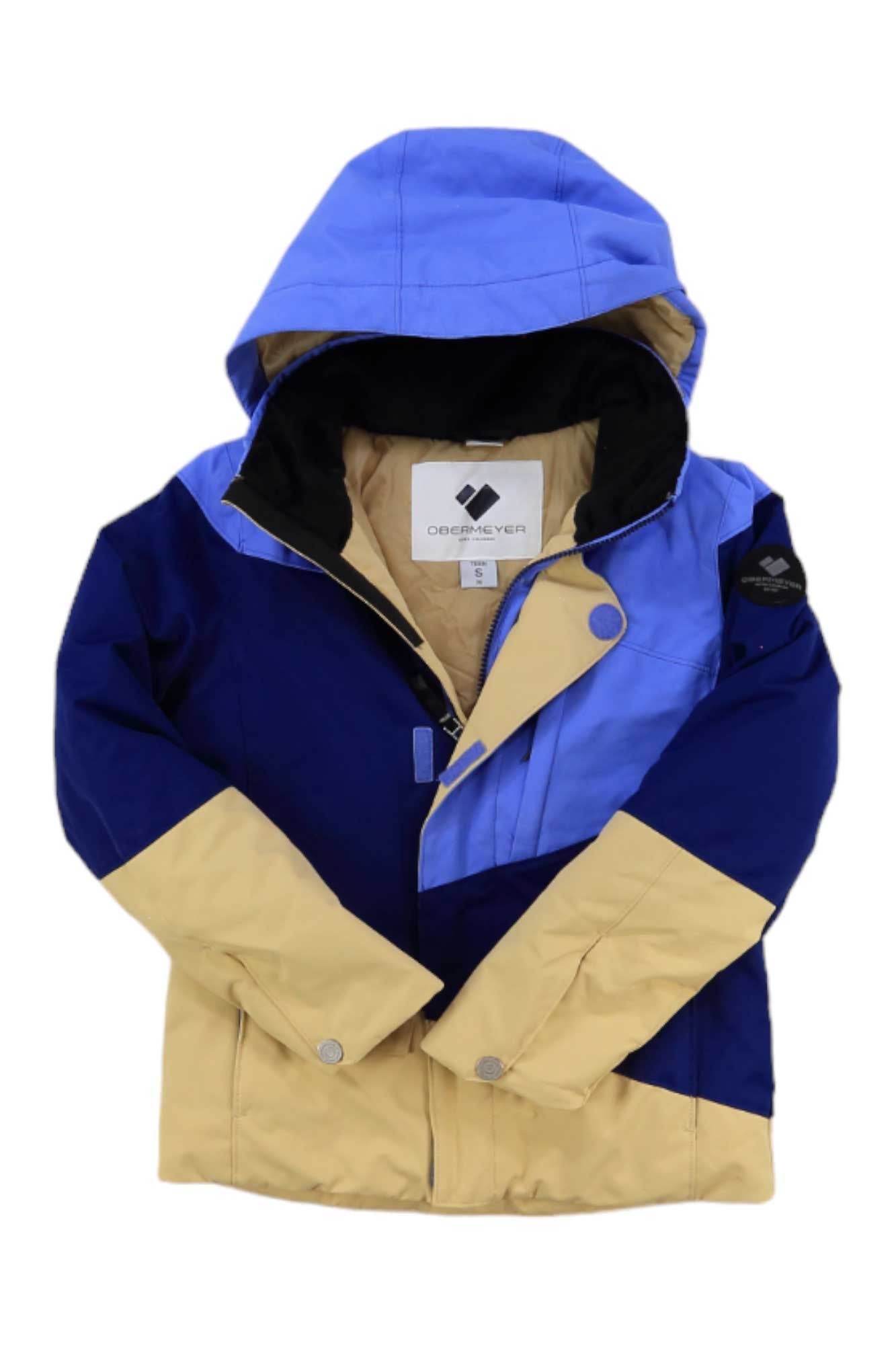 The Obermeyer Girls Taylor Jacket is a color-blocked kids’ hooded jacket with beige, dark blue, and light blue panels, front zipper and snap closures. The black interior features a visible Obermeyer brand tag.