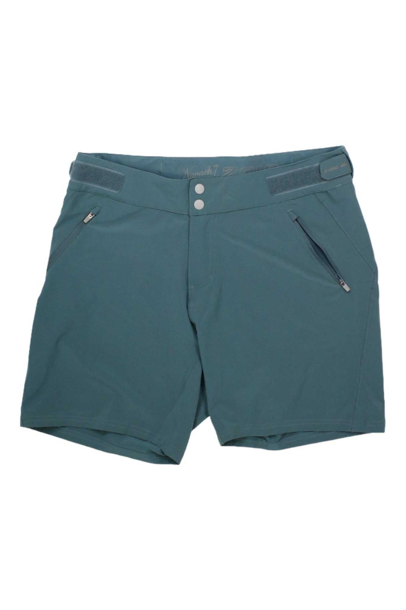 The Zoic Womens Navaeh 7 Short in teal is a knee-length cycling short featuring front zipper pockets, belt loops, and a double snap button closure, shown laid flat on a white background.
