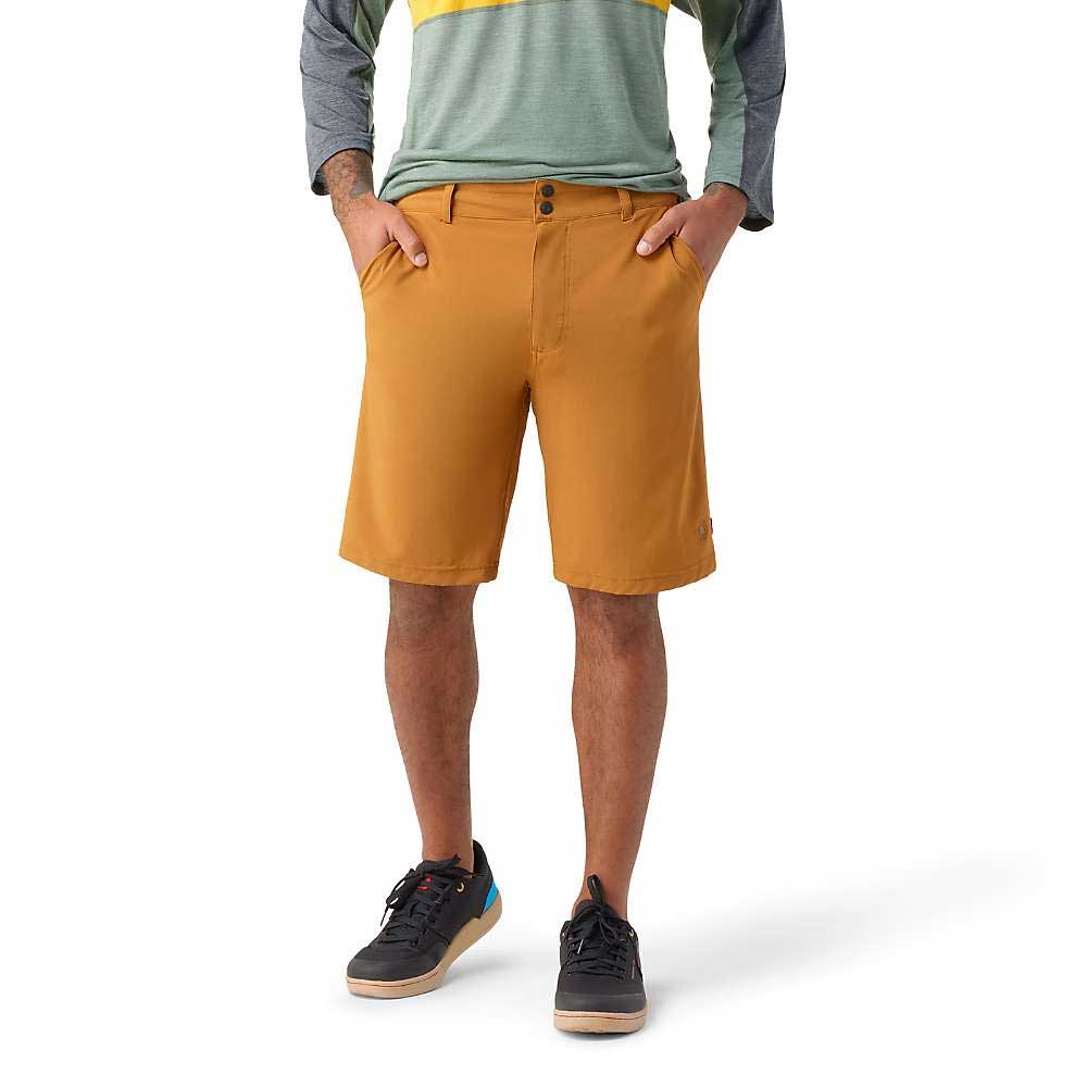A person stands with hands in pockets, wearing mustard-colored Smartwool Mens 10 Inch Short, a long-sleeve green and gray shirt, and black sneakers with white soles against a plain white background.