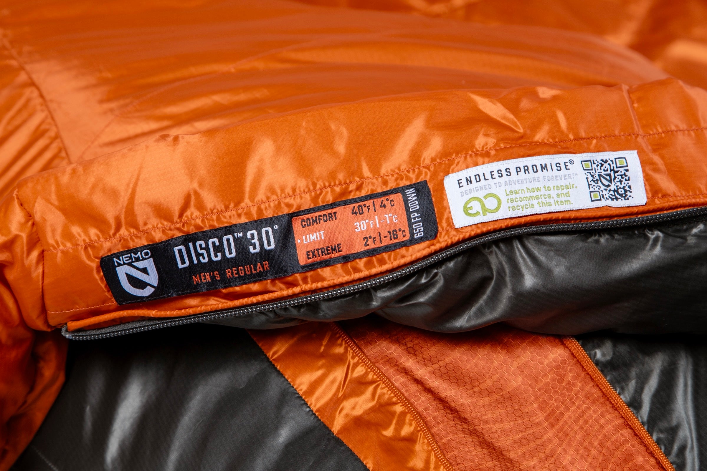 Close-up of the Nemo Mens Disco 30 Sleeping Bag (orange/gray, 3-season), showing its label with temperature ratings, brand logo, QR code, and recycling info; model: DISCO 30° MENS REGULAR—designed for side-sleepers.