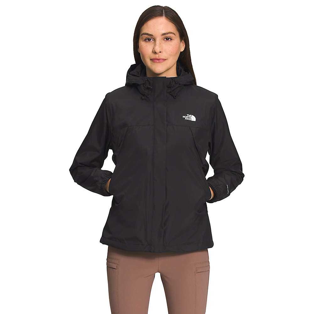 A woman with straight brown hair faces forward, wearing The North Face Womens Antora Triclimate Jacket in black with her hands in the pockets, paired with light brown pants, against a plain white background.