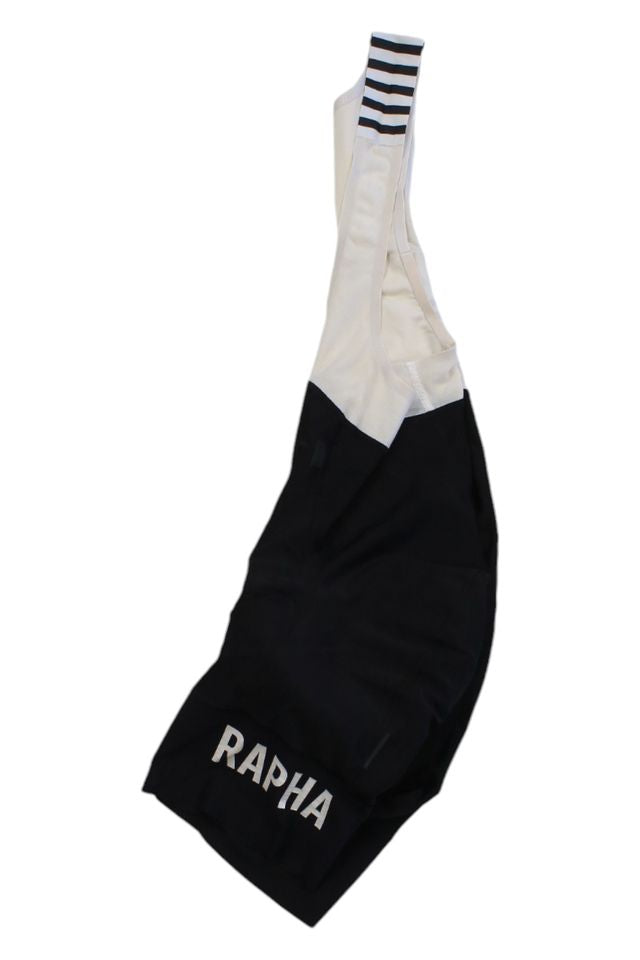 Black and white Rapha Mens Pro Team Bib Shorts II - Regular, featuring bold white RAPHA lettering on the leg, black stripes on one shoulder strap, and a race-grade chamois, displayed against a white background.