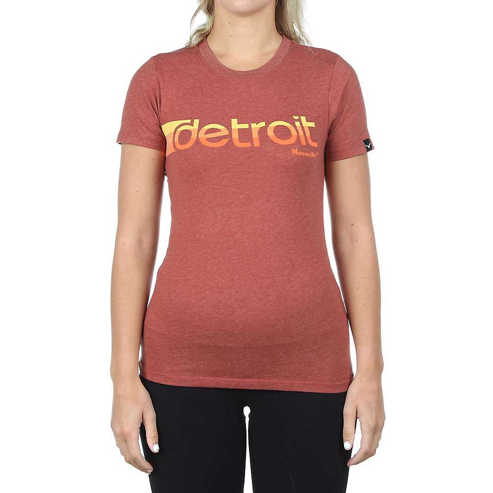 A woman wears a fitted Moosejaw Women's Retro Axel Foley SS Tee—red with "detroit" in bold yellow-orange letters—paired with black pants, standing against a white background.
