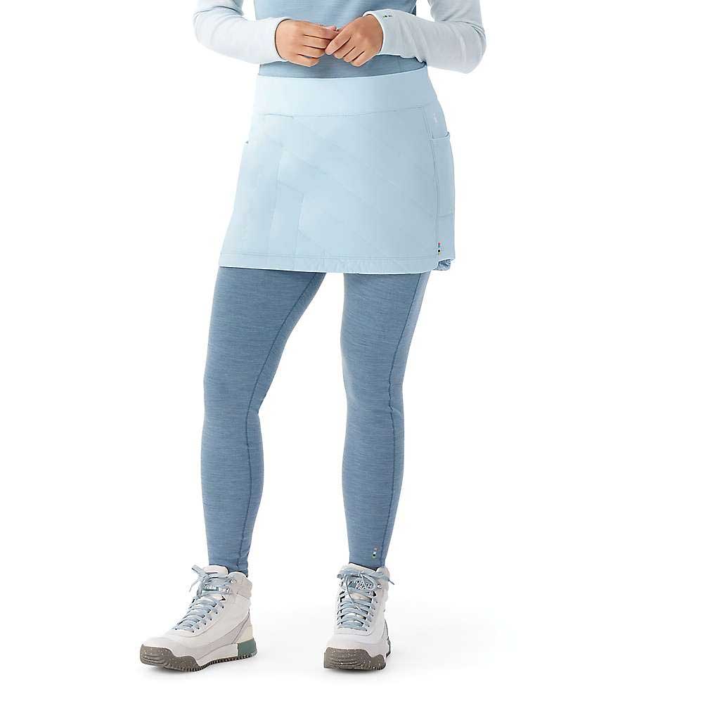 A person stands against a white background, wearing a light blue long-sleeve top, Smartwool Women's Smartloft Skirt, blue leggings, and white hiking shoes, with hands resting near their waist.