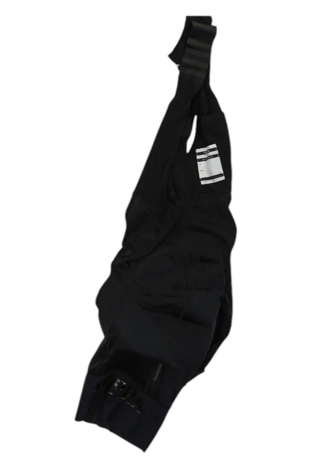 A pair of Rapha Mens Pro Team Bib Shorts II - Regular with shoulder straps and a small white tag, featuring a race-grade chamois, laid flat on a white background.