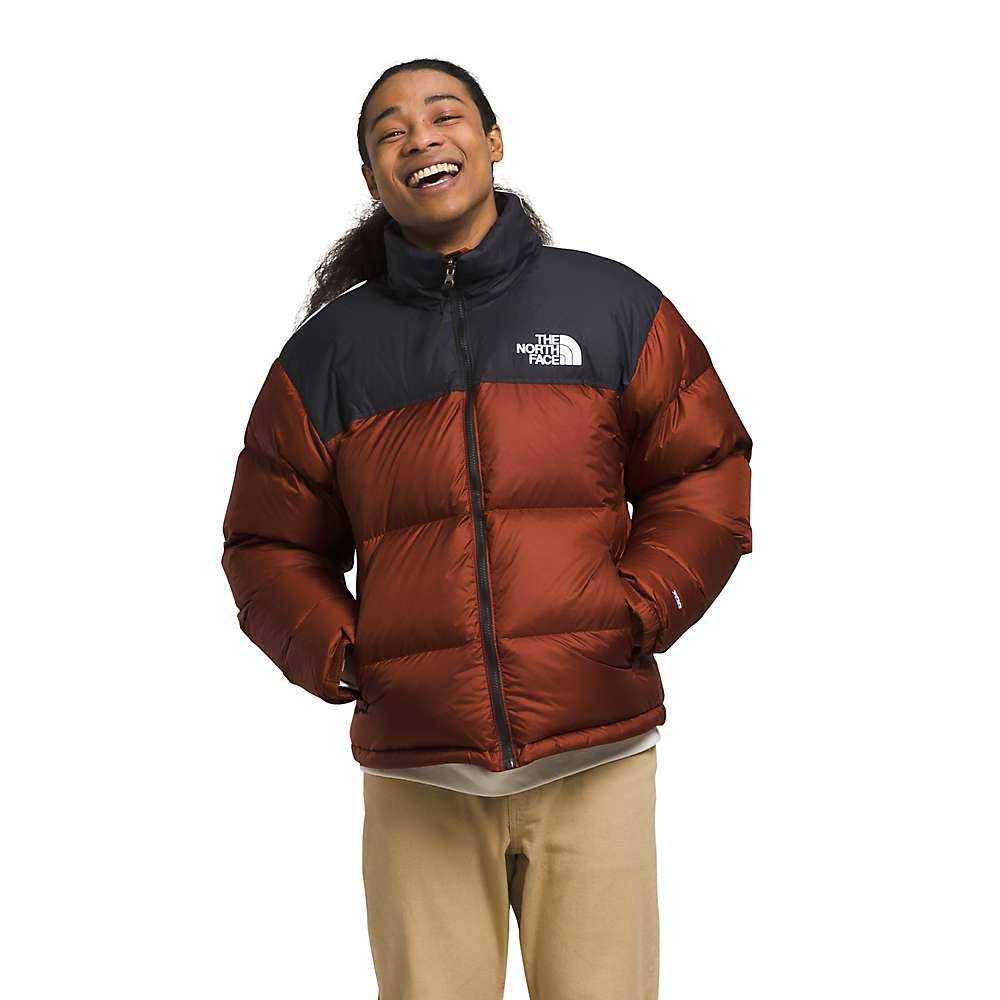 A person smiles with hands in pockets, wearing The North Face Men's 1996 Retro Nuptse Jacket in shiny red and black with 700-fill down insulation, beige pants, and a cream shirt, standing against a plain white background.