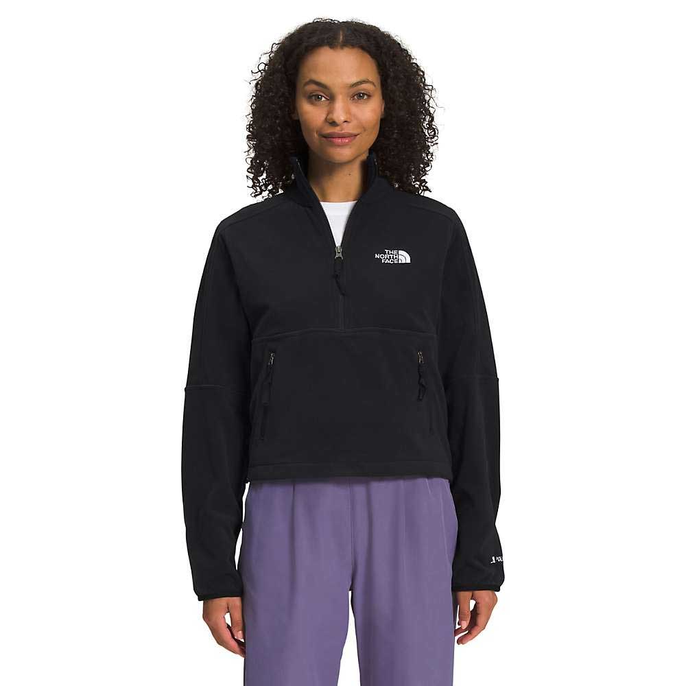 A woman with curly hair faces the camera wearing The North Face Women's TNF Polartec 100 1/4 Zip Jacket in black with long sleeves and loose purple pants. The jacket is made from recycled fleece.