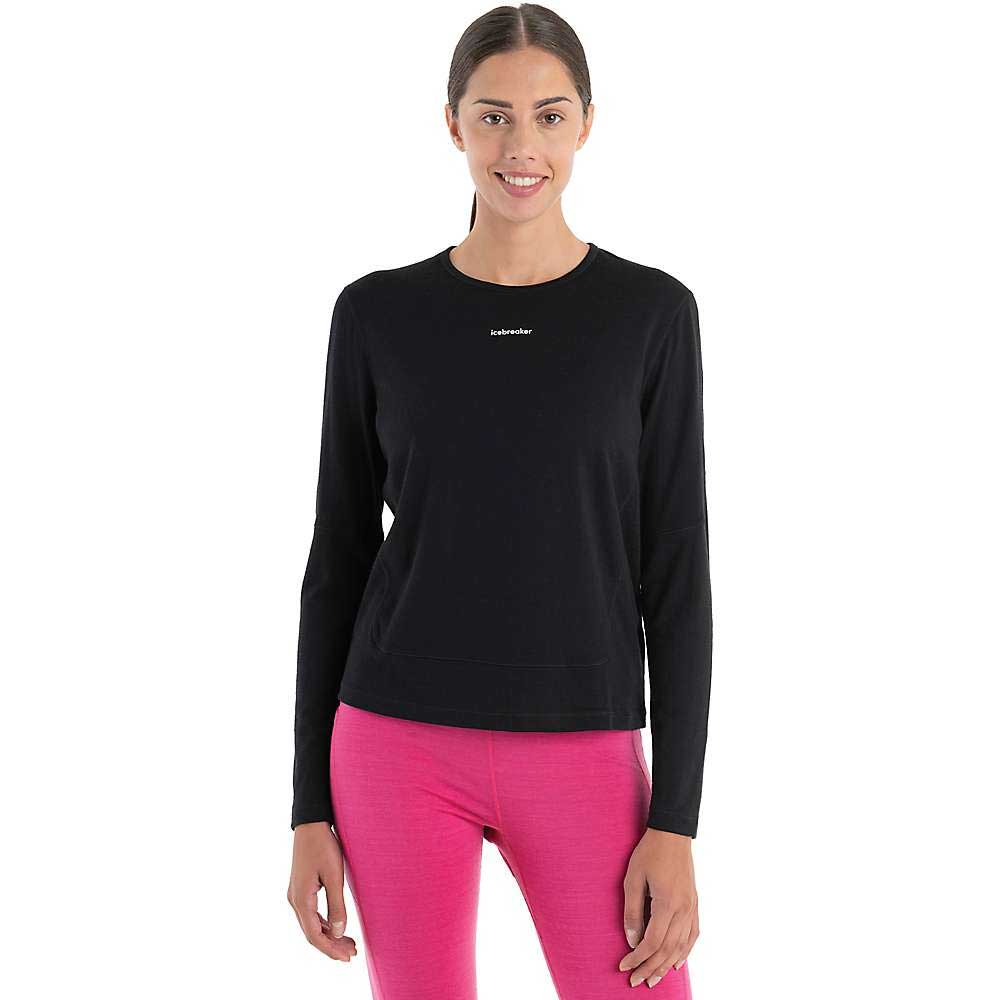 A woman with brown hair tied back is smiling and wearing the Icebreaker Womens Merino 200 ZoneKnit Energy Wind LS Tee in black with white text, paired with bright pink pants, standing against a plain white background.