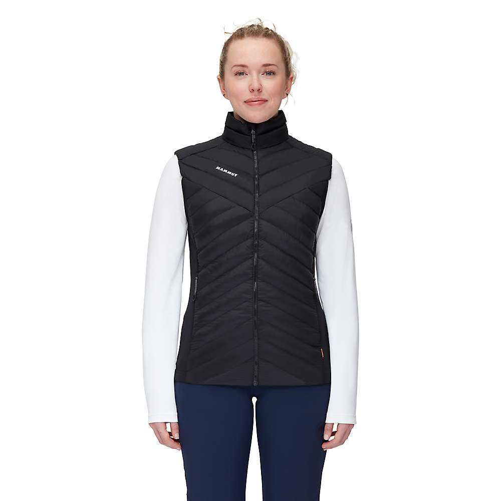 A person stands facing forward, wearing the Mammut Womens Albula IN Hybrid Vest—black with recycled synthetic insulation—over a white long-sleeve shirt and dark pants, set against a plain white background.