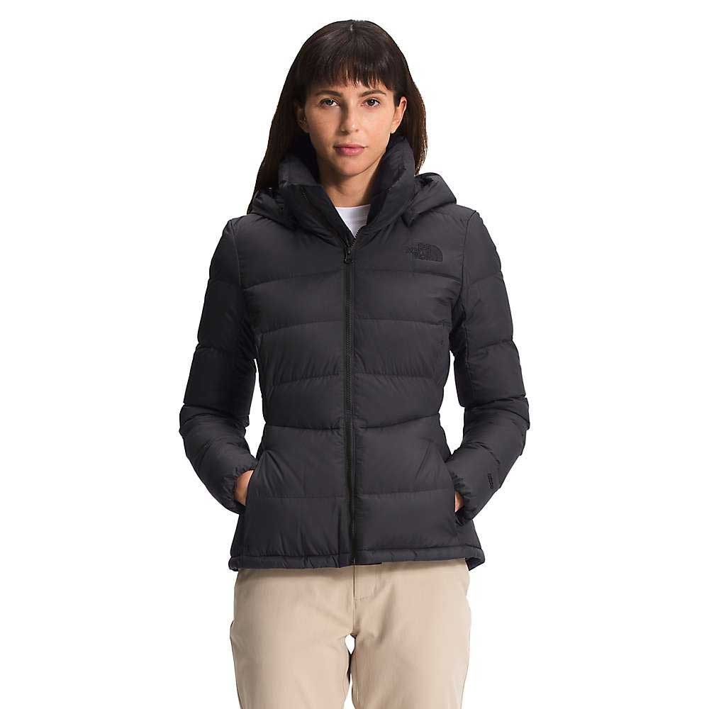 A woman with straight dark hair and bangs wears The North Face Womens Metropolis Jacket, a 550 fill goose down jacket with a hood, pairing it with light pants. She stands against a white background with her hands in the pockets.
