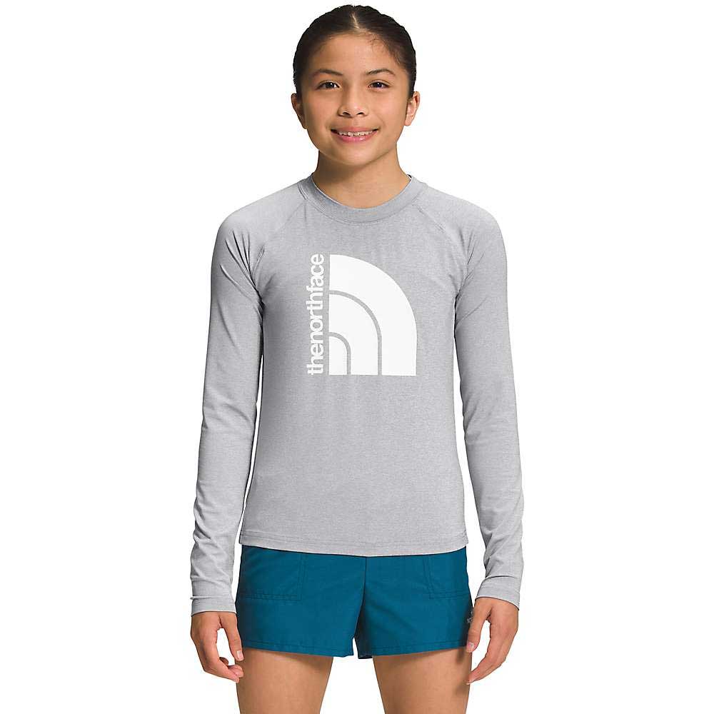 A girl stands smiling in a light gray The North Face Girls Amphibious LS Sun Tee with a large white logo on the front and teal shorts, set against a plain white background.