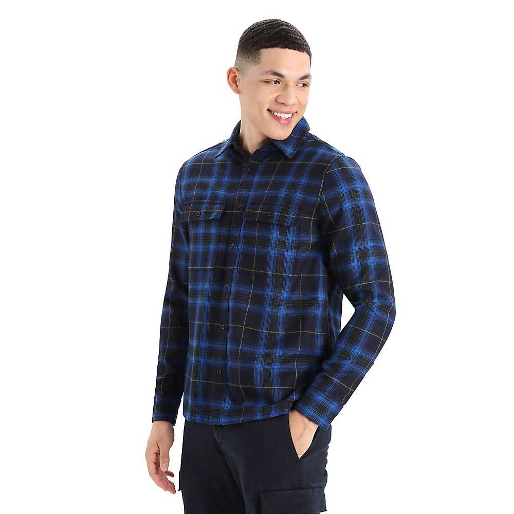 A young man smiles in the Icebreaker Mens Dawnder LS Flannel Shirt Plaid, blue and black, paired with black pants, set against a plain white background.