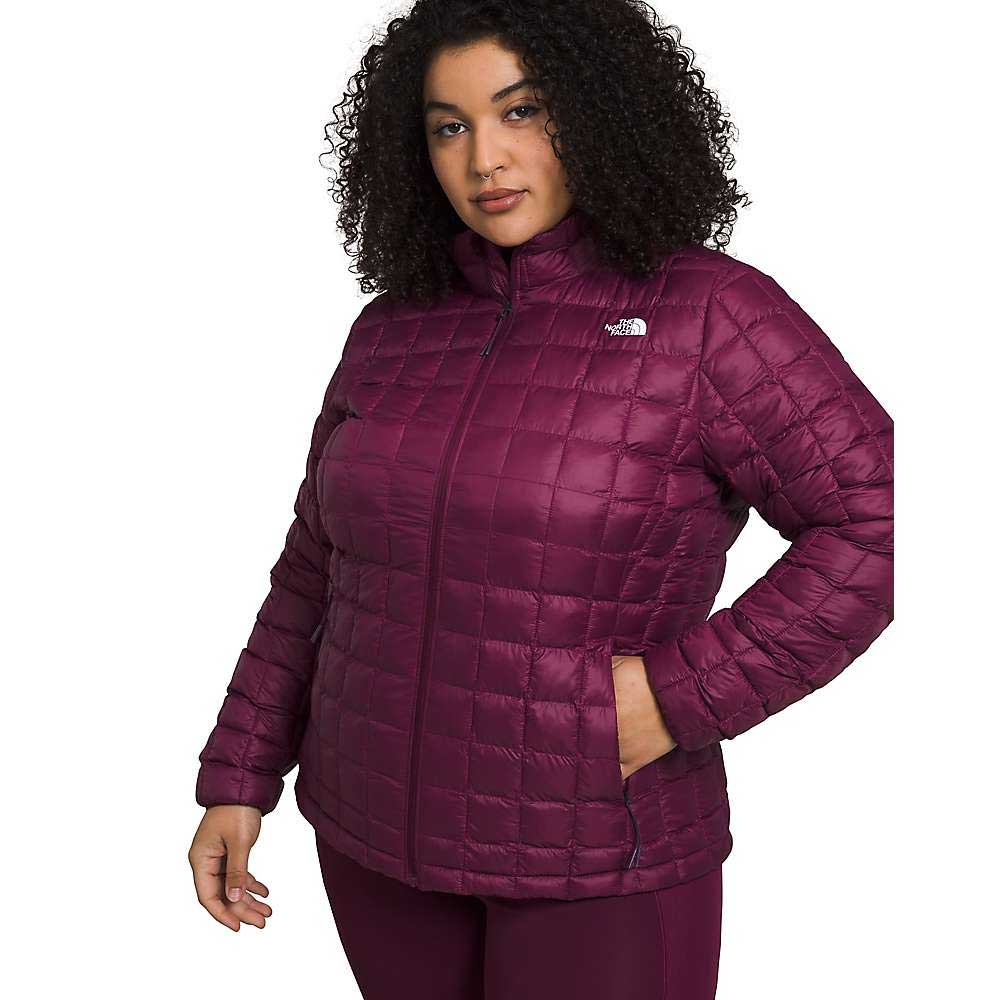 A woman with curly hair wears The North Face Womens Plus ThermoBall Eco 2.0 Jacket in quilted dark magenta, made from recycled nylon, hands in the pockets, standing against a plain white background.