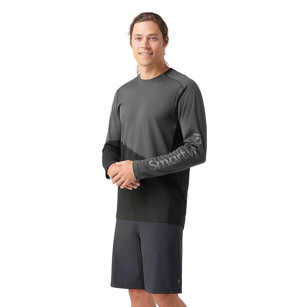 A person with long hair in a ponytail smiles while wearing a moisture-wicking, dark gray Smartwool Mens Mountain Bike LS Jersey and matching shorts, standing against a white background.