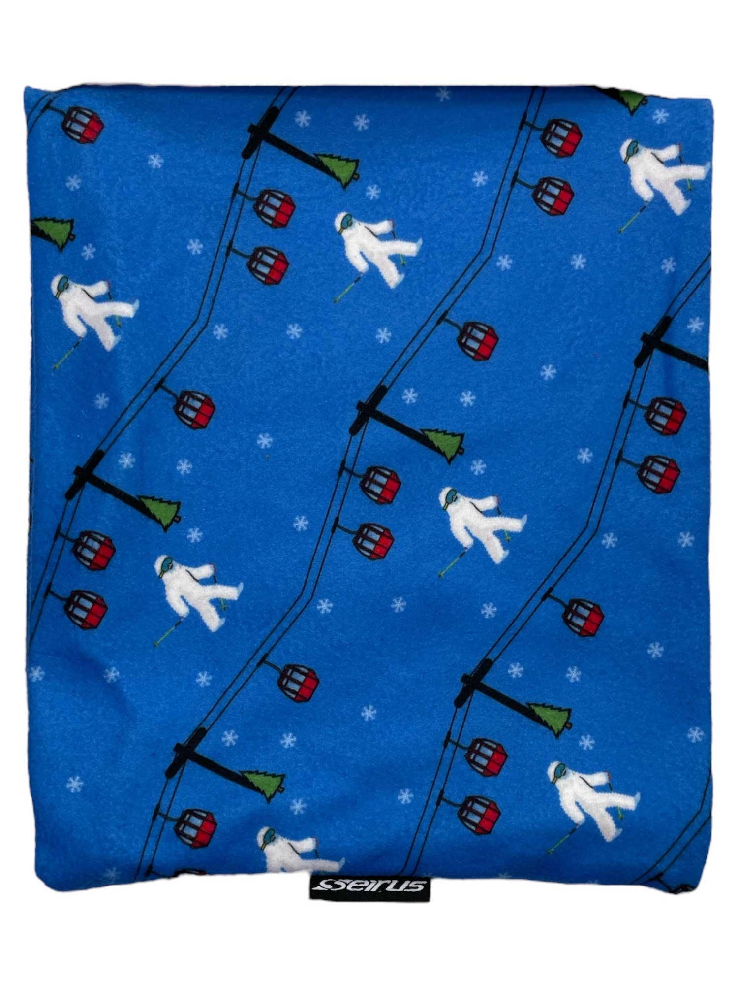 The Seirus Junior Micro Fleece Neck Up Gaiter is a blue kids' winter neck warmer featuring white skiers, red gondolas, green pine trees, tiny snowflakes, and a visible Seirus brand tag at the bottom.