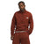 A man in a rust-red The North Face Men's Canyonlands 1/2 Zip Jacket and matching pants stands with one hand in his pocket, looking at the camera against a plain white background.