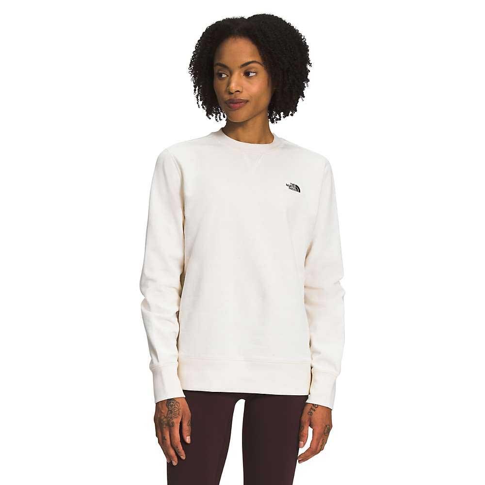 A woman, wearing The North Face Womens Heritage Patch Crew in light beige—crafted from recycled fabric—and dark pants, stands facing forward with hands at her sides, gazing slightly to the left.