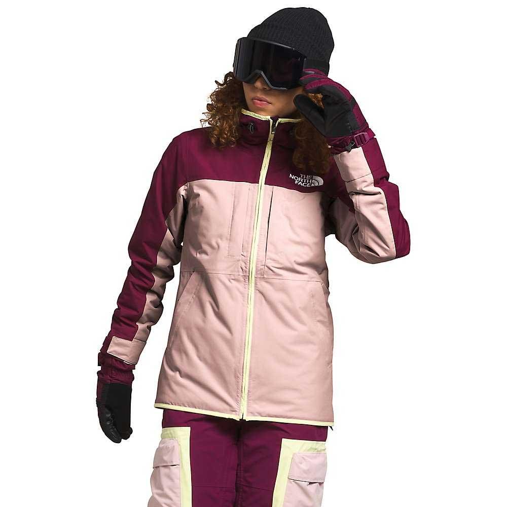 A person wears a maroon and light pink The North Face Womens Namak Insulated Jacket with Heatseeker Eco, matching pants, black gloves, dark ski goggles, and a black beanie, standing against a white background.