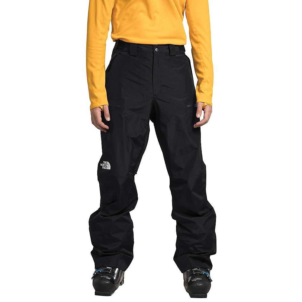 A person wearing a yellow long-sleeve shirt, The North Face Mens Dawnstrike GTX Pant in black, and black ski boots stands facing forward against a white background.