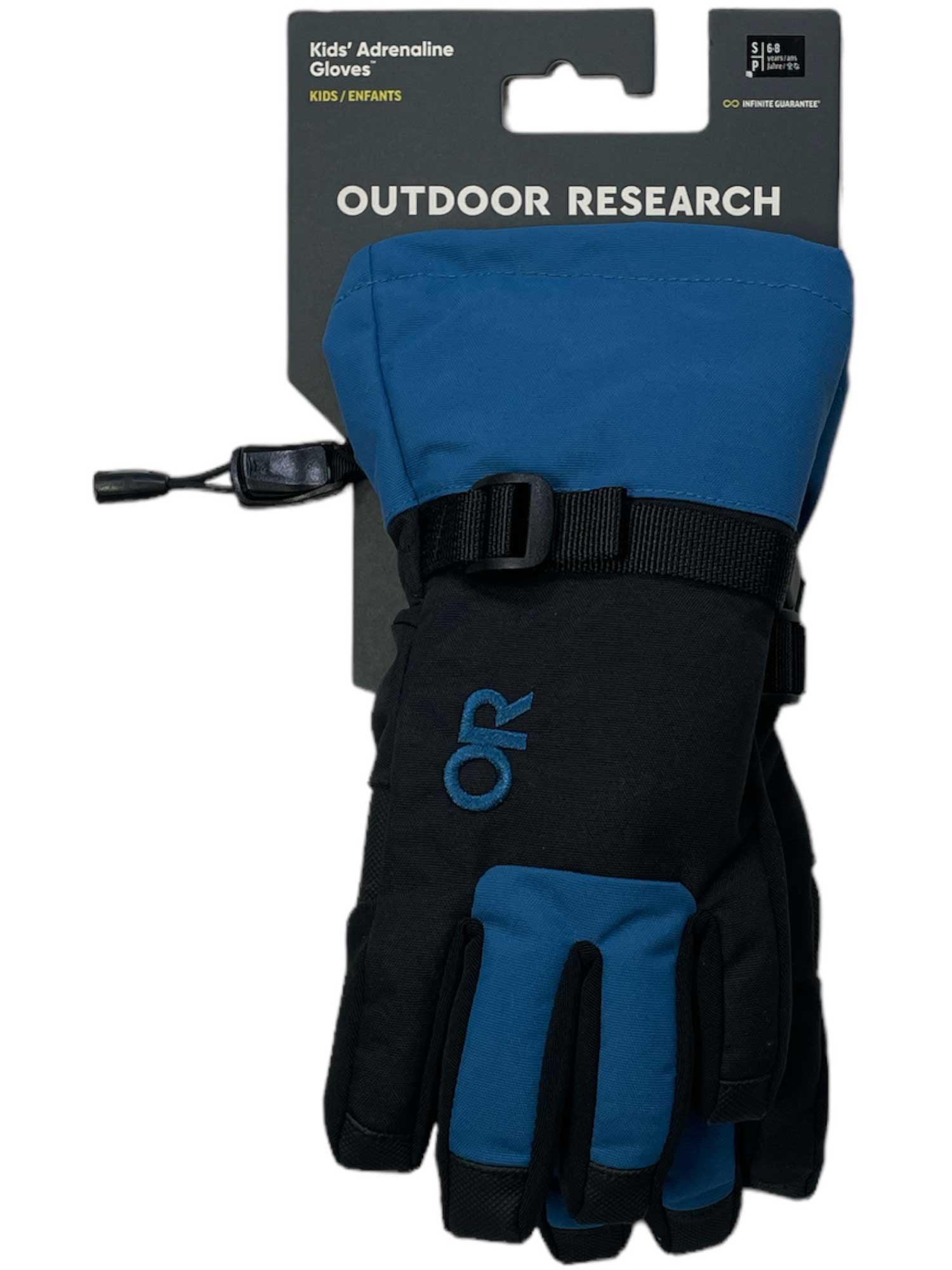 A pair of blue and black Outdoor Research Kids Adrenaline Glove winter gloves with a strap, adjustable wrist buckle, “OR” logo on the back, and attached to black packaging.