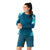 A woman stands smiling with her arms loosely crossed, wearing the Smartwool Women's Mountain Bike LS Jersey in a blue and teal pattern and matching teal shorts.