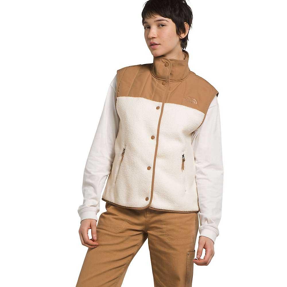 A woman wears The North Face Women's Cragmont Fleece Vest in tan and white over a white long-sleeve shirt and tan pants, standing with one hand in her pocket against a plain background.