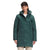A woman with straight brown hair wears The North Face Womens Arctic Triclimate Jacket in dark green, paired with light pants and a white shirt, standing facing forward with a neutral expression.