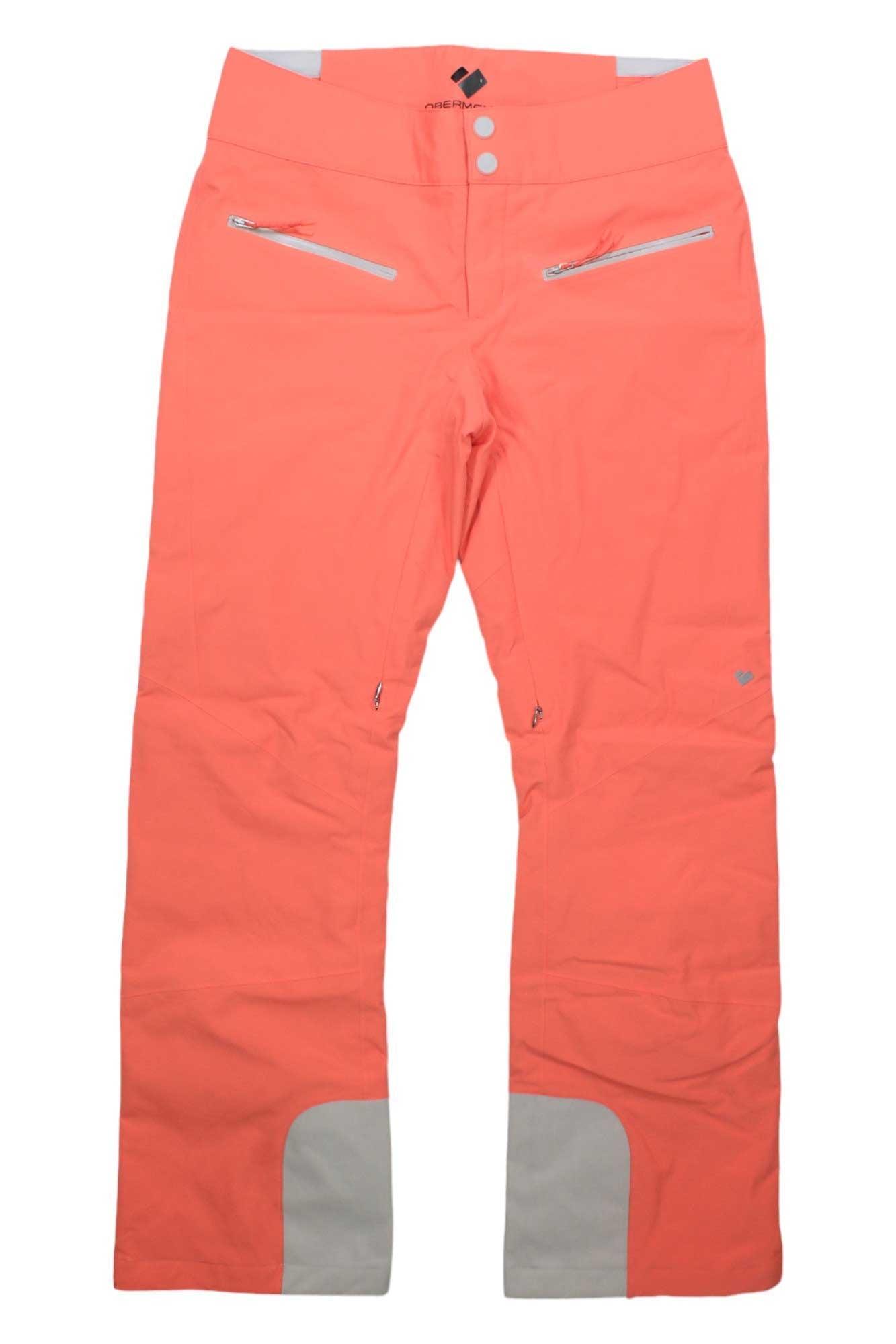 The Obermeyer Womens Bliss Pant features a coral color, two front zip pockets, white waistband accents, and light gray reinforced inner ankle patches. These insulated ski pants are ideal for outdoor or athletic activities.