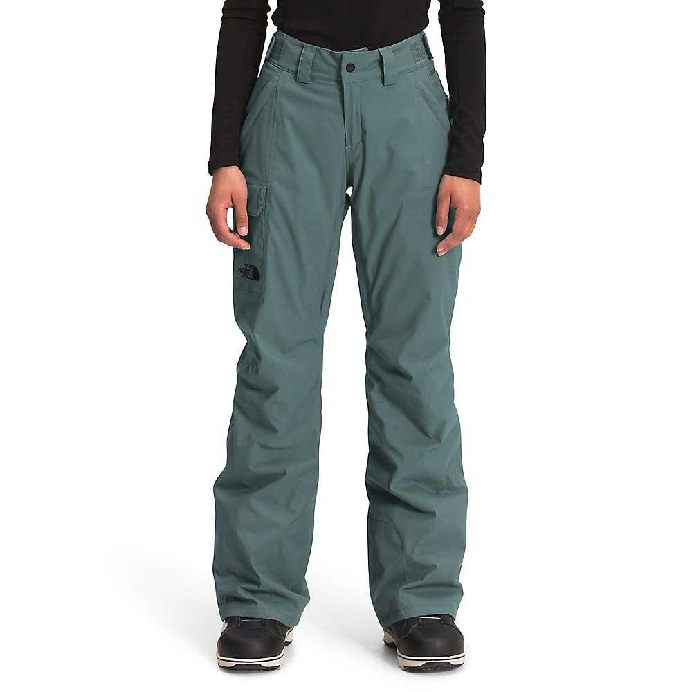 A person wearing teal green The North Face Womens Freedom Insulated Pant, paired with a black long-sleeve top and black boots, stands against a white background.