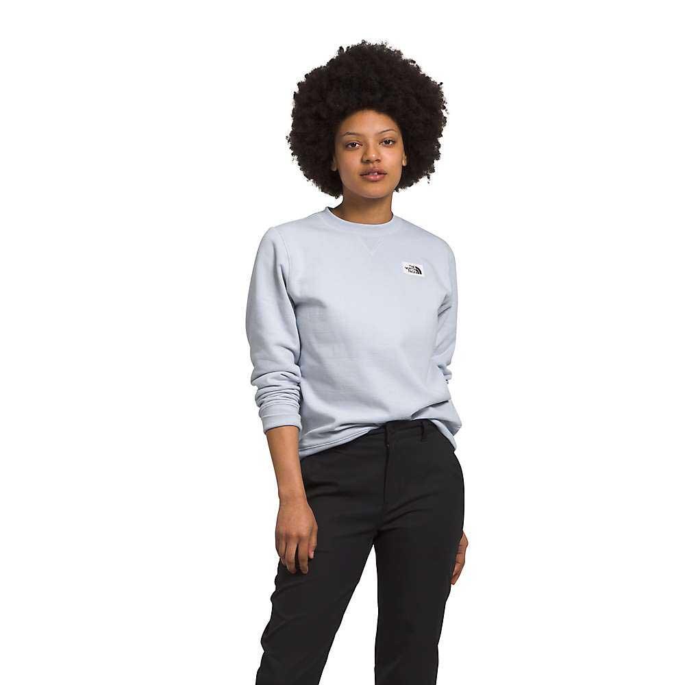 A woman with short curly hair wears The North Face Womens Heritage Patch Crew, a recycled-fabric sweatshirt, with black pants against a plain white background.
