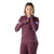 A woman with curly hair wears a maroon Smartwool Women's Intraknit Merino Tech 1/4 Zip Top with light purple sleeve stripes and matching pants, posing with one hand near her chin and the other resting on her sleeve.