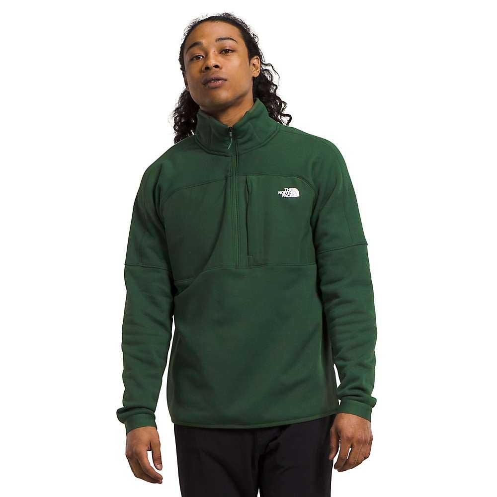 A person wearing The North Face Mens Canyonlands High Altitude 1/2 Zip Top in dark green, made from recycled polyester with a zippered chest pocket, stands against a plain white background.