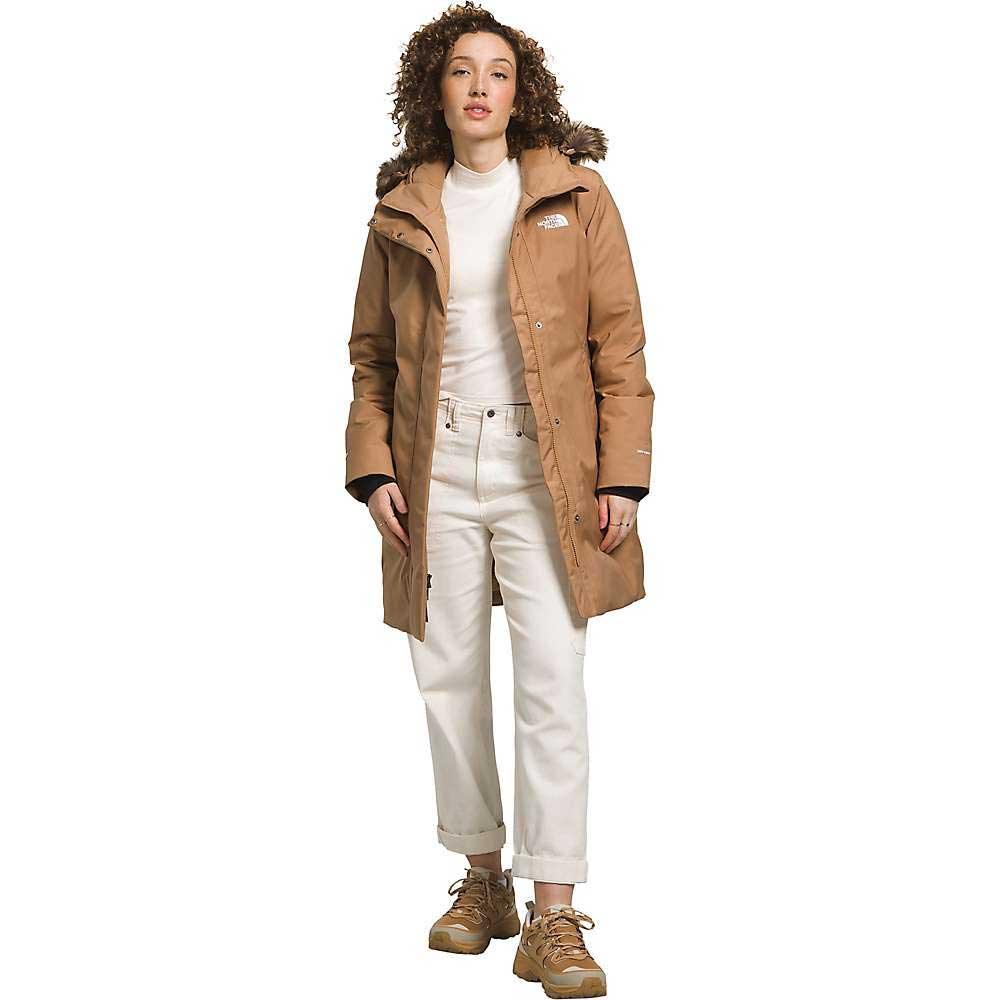 A woman with curly hair wears The North Face Womens Arctic Parka in tan over a white turtleneck, paired with white cuffed jeans and tan sneakers, standing against a plain white background.