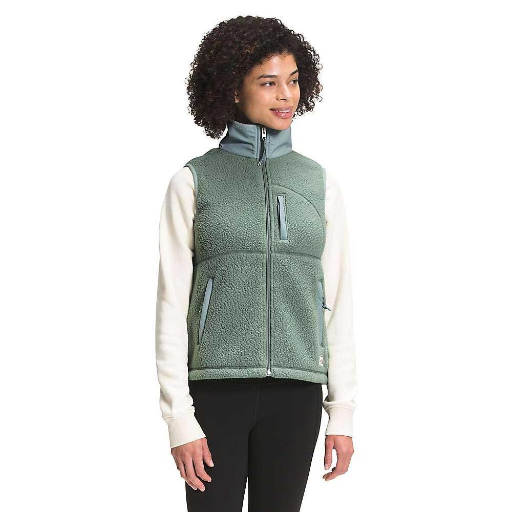 A woman with curly hair wears The North Face Women's Cragmont Fleece Vest in sage green over a cream long-sleeve shirt and black pants, standing and smiling slightly while looking to the side.