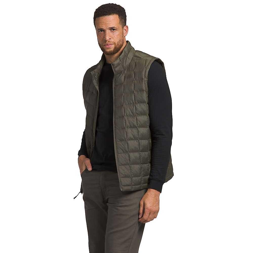 A man poses against a white background wearing The North Face Men's Big ThermoBall Eco 2.0 Vest in olive green over a black long-sleeve shirt and gray pants, with one hand in his pocket and a neutral expression.