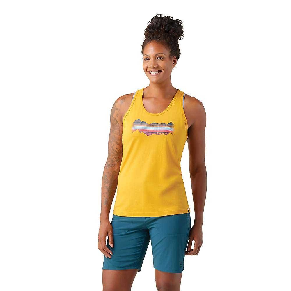 A woman with curly hair in a bun smiles while wearing the Smartwool Women's Mountain Horizon Graphic Tank and teal shorts, standing against a plain white background.