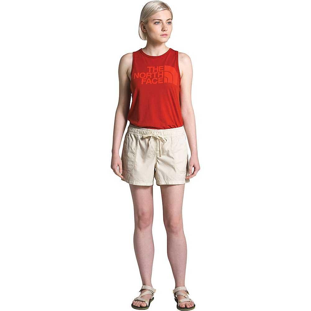 A woman with light skin and blonde hair faces forward, wearing a red sleeveless tank, The North Face Women's Motion Pull-On 4 Inch Shorts in beige, and beige sandals. She gazes slightly to the side.