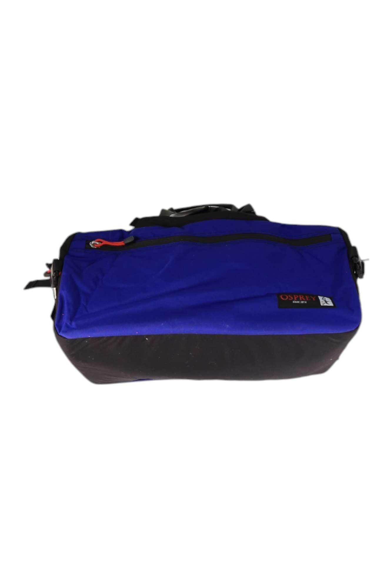 The Osprey Heritage Transporter 45 Duffle Bag is rectangular, made from recycled nylon with a blue top, black bottom, front zipper pocket, sturdy handles, and a logo patch on the front.