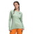 A woman with braided hair models The North Face Womens Fd Pro 160 Crew in light green, made from recycled polyester, paired with bright orange pants. She stands with one hand on her hip against a plain white background.