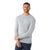 A man with short curly hair and a beard stands smiling in a plain white setting. He wears a light gray Smartwool Mens Sparwood Crew Sweater with dark blue jeans, arms crossed while his other hand holds his wrist.