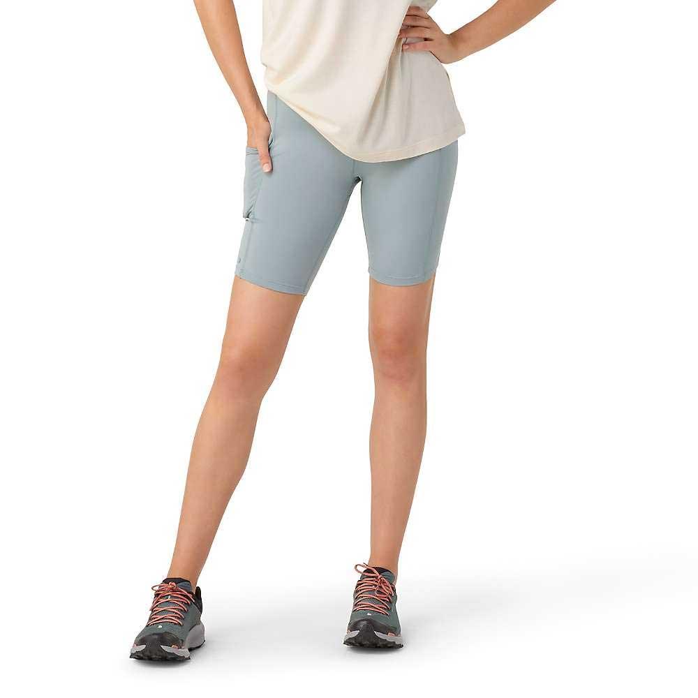 Wearing a white shirt, Smartwool Women's Active Biker Short in light blue with a high-rise waist and side pocket, and black athletic shoes with pink laces, a person stands with one hand on their hip against a white background.